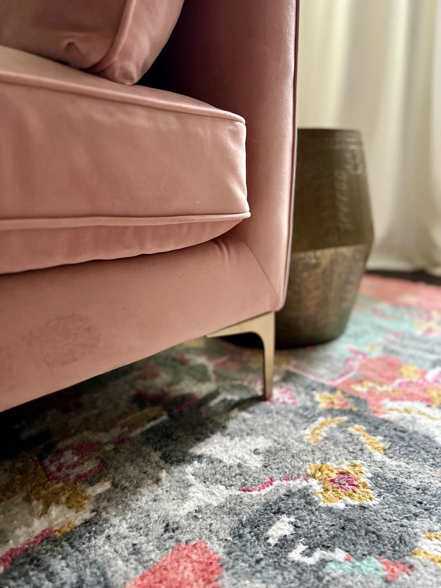 Close-up of a dusty rose-colored sofa corner with tapered legs on a colorful textured rug, next to a brass-colored textured side table.