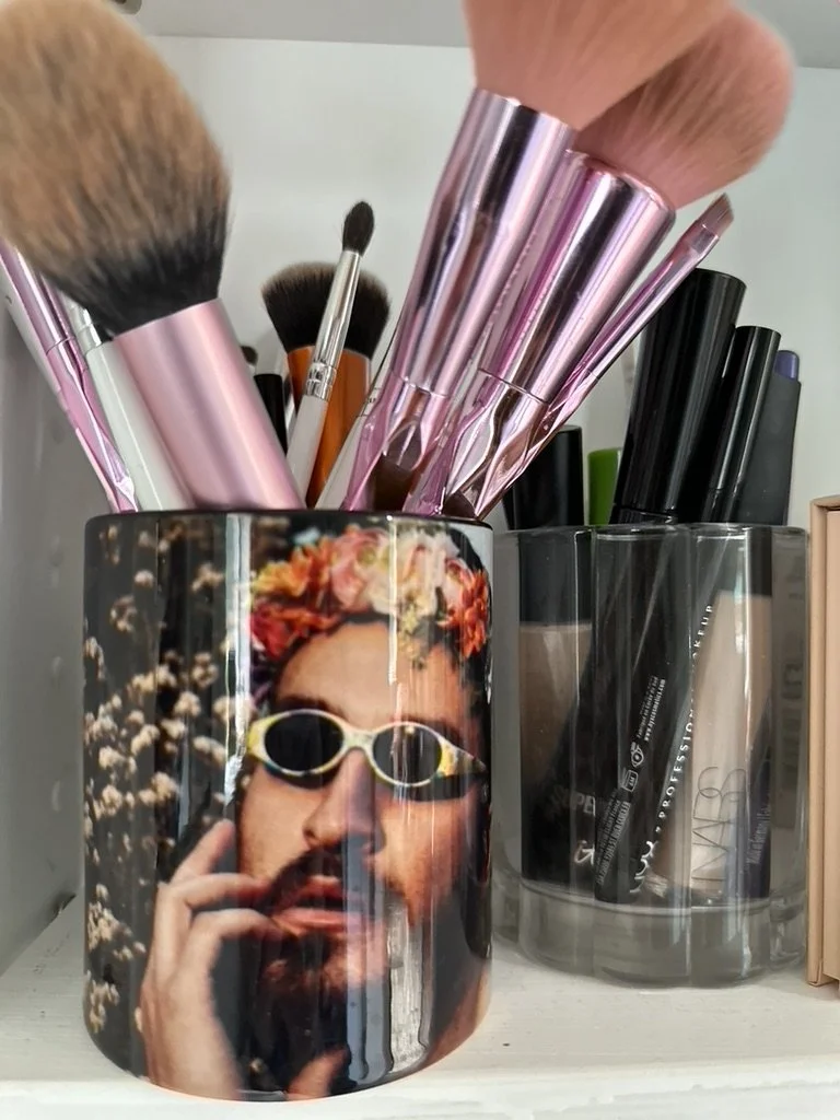 Makeup brushes in a mug with a print of a person wearing sunglasses and a floral crown, next to a container of pens and makeup products on a shelf.