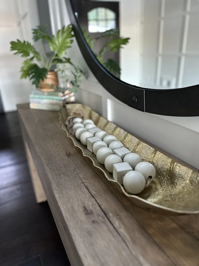 A decorative tray on a wooden console table holding white spherical and cubic objects, accompanied by potted plants and stacked books. A round mirror is mounted on the wall above.
