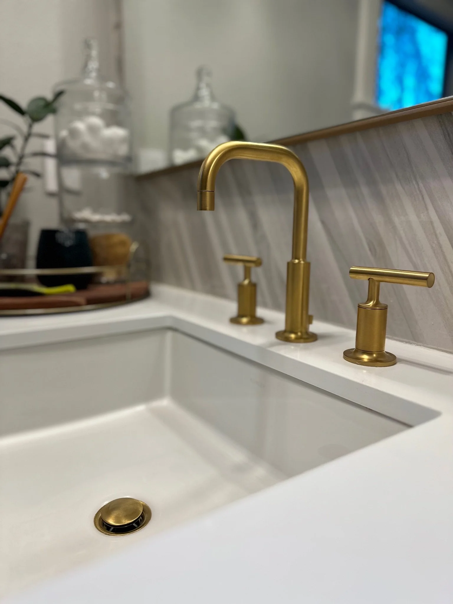 Bathroom sink with gold faucet and handles, white countertop, and decorative glass jars in the background.