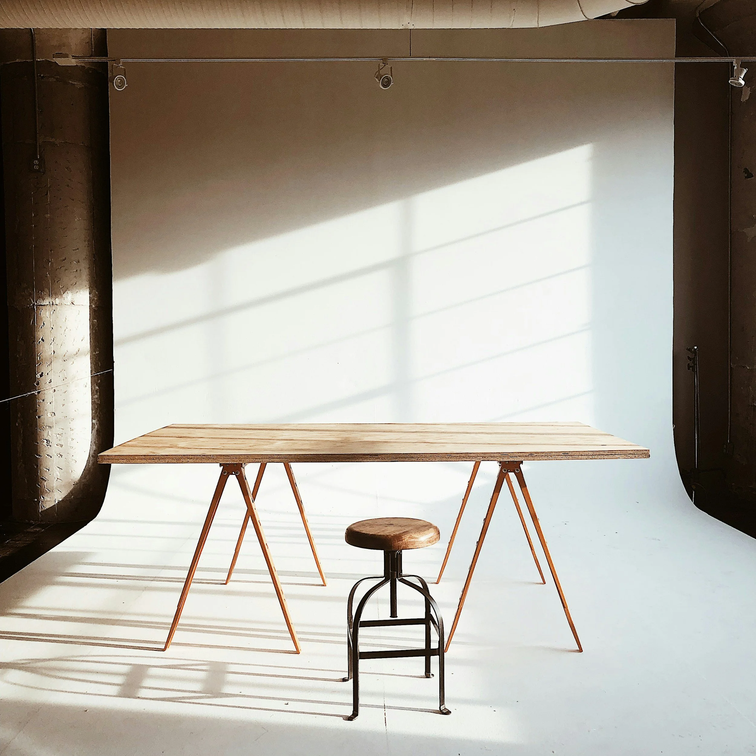 A wooden table with angled legs and a matching wooden stool with metal legs, set against a plain white wall with shadows cast by window blinds.