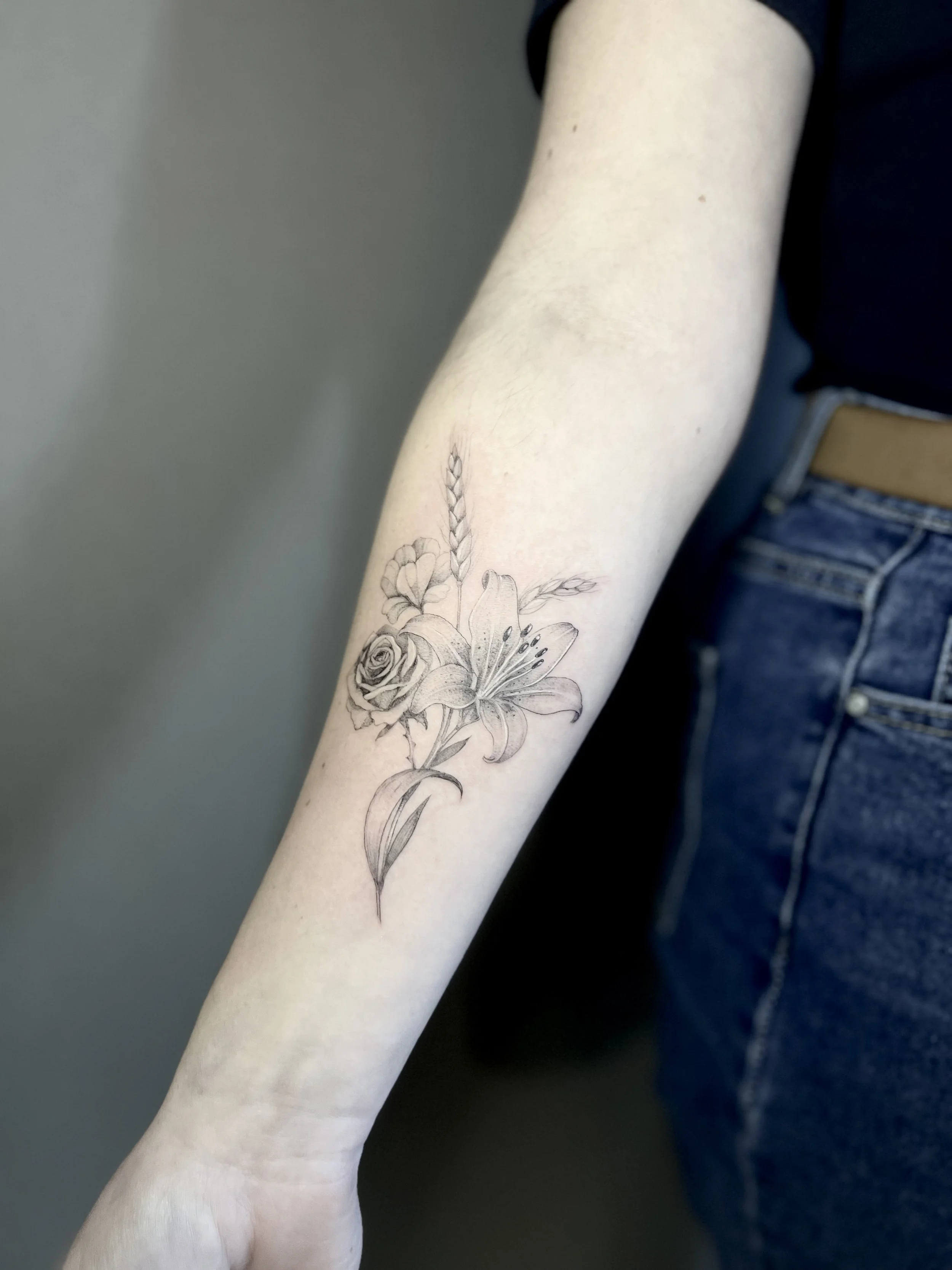 Tatouage d'un bouquet de fleurs comprenant une rose, un lys, un épi de blé et une feuille, en style de dessin au trait noir.