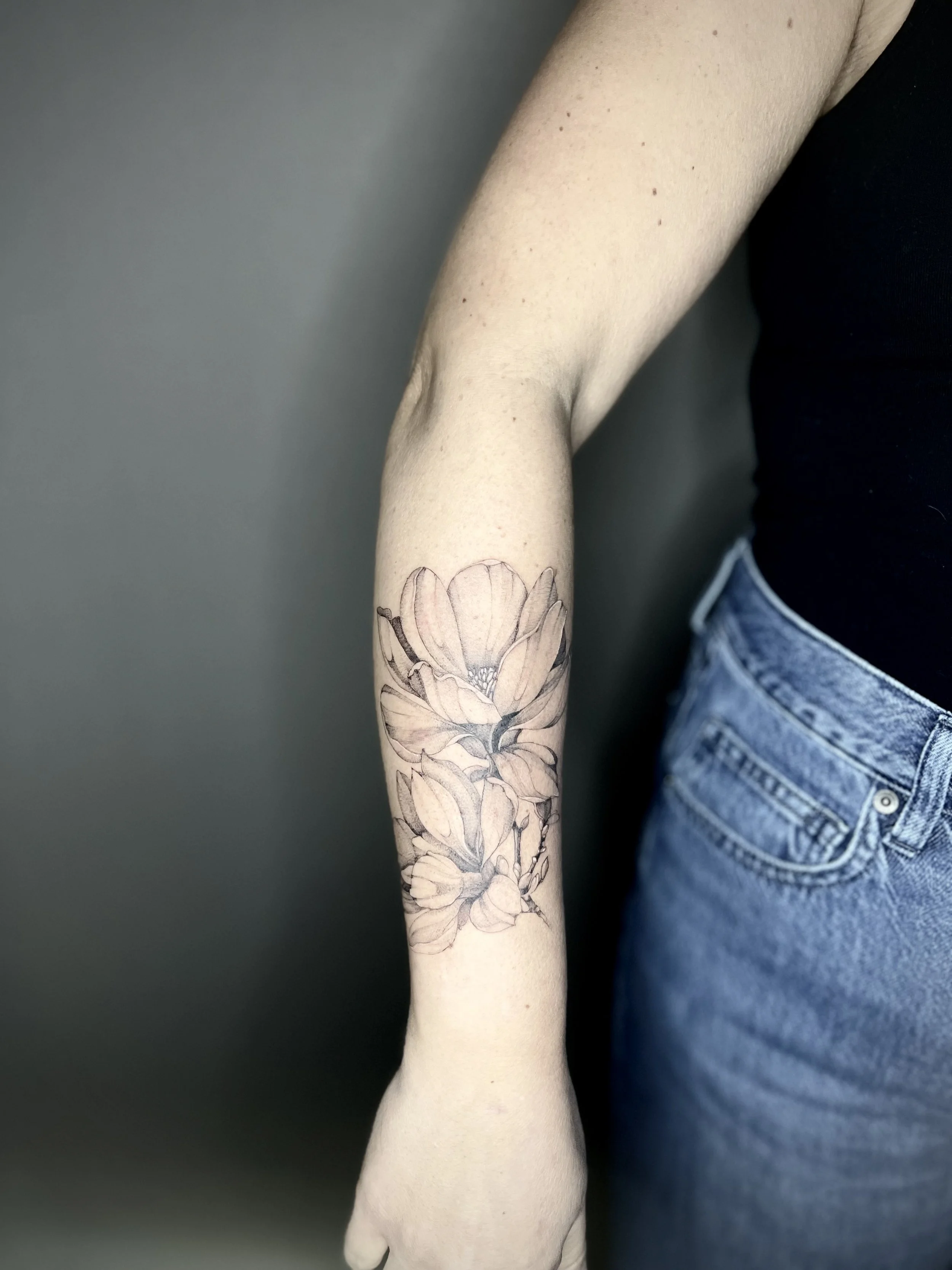 Tatouage de fleurs sur le bras d'une personne, avec un fond gris et un t-shirt noir, portant un jean.