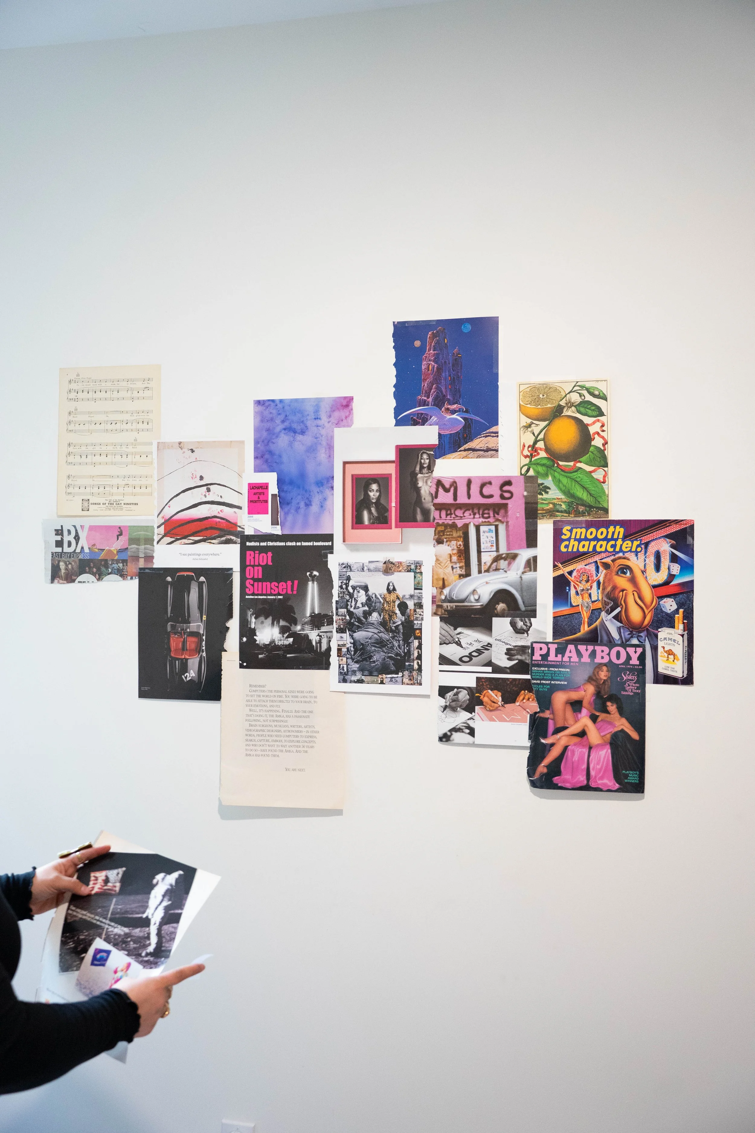 Person holding printed photographs in front of a white wall decorated with various posters, photos, and artworks.