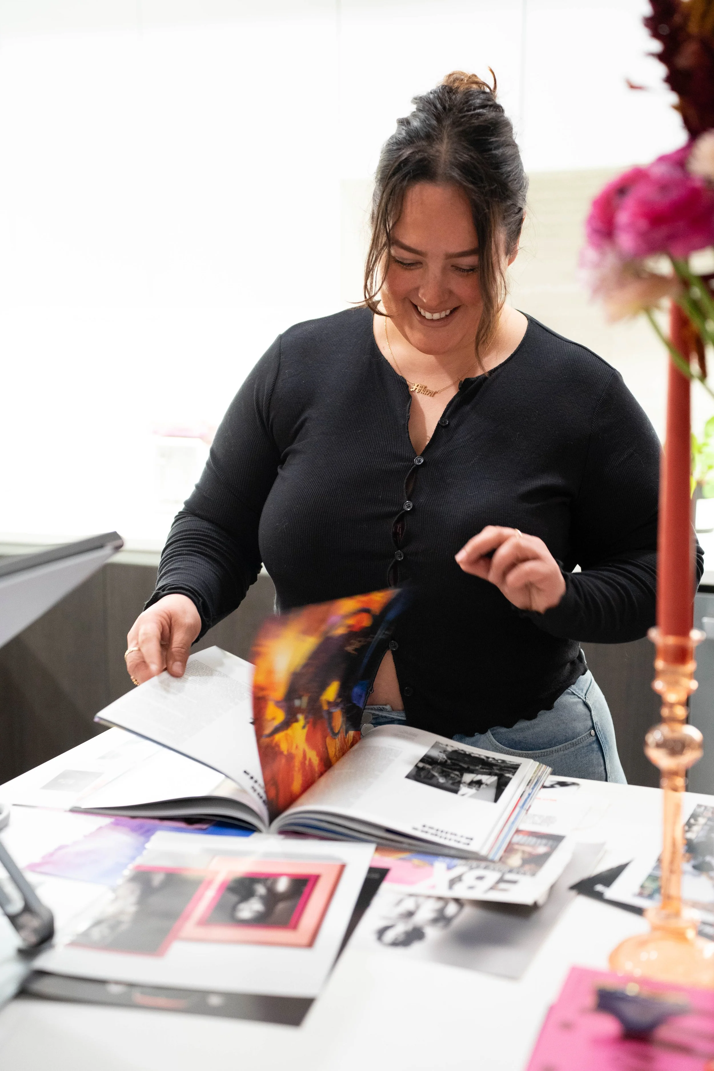 A woman in a black top smiling and looking down as she flips through a large magazine or photo book on a table, with various photographs and images spread out on the table.