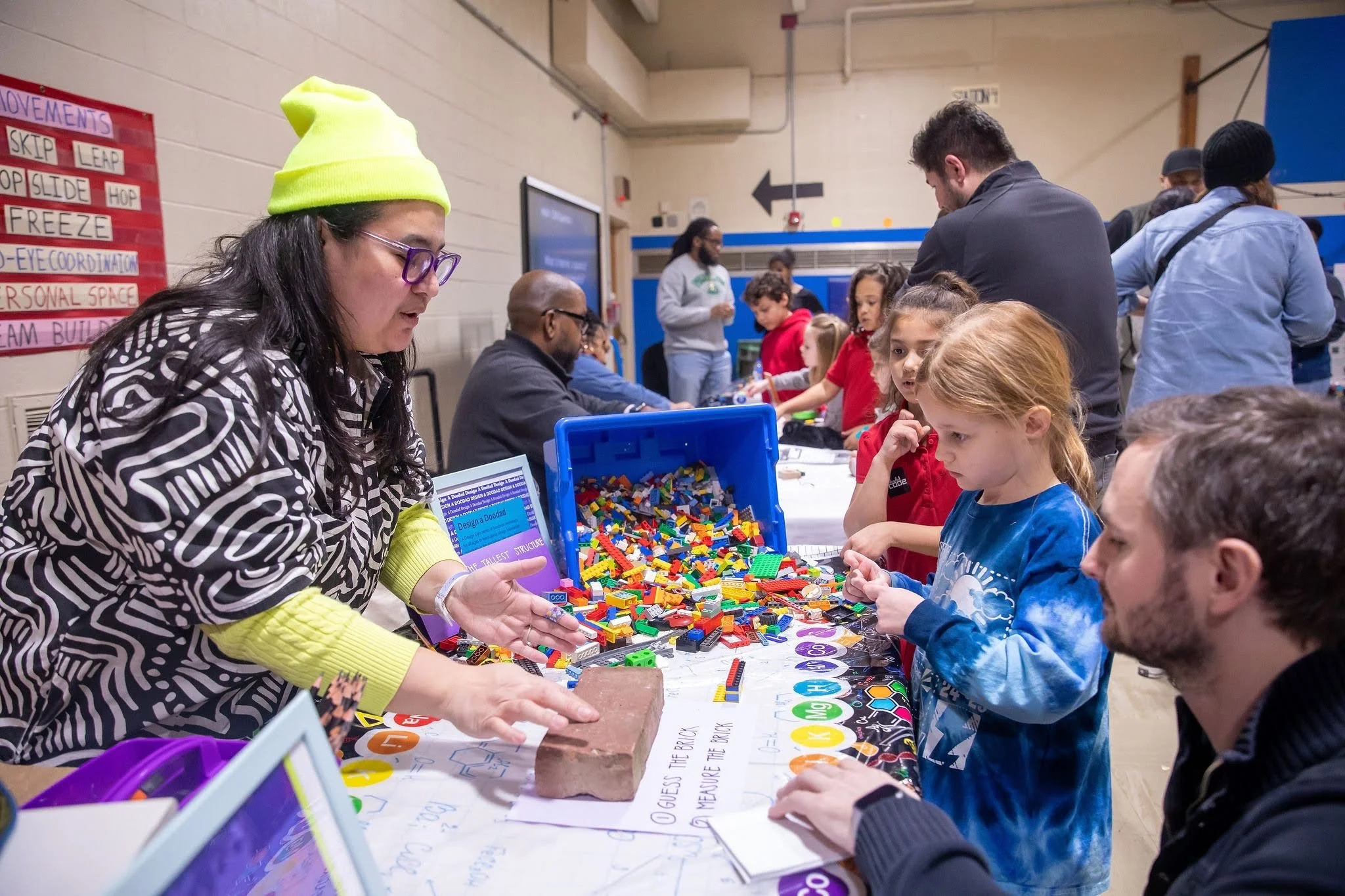 Erike, founder of Design Gym, is asking young scholars if they can guess the dimensions of a single red brick. Beyond the brick is a rectangle container of Lego bricks on its side with Lego bricks spilling over onto the table.