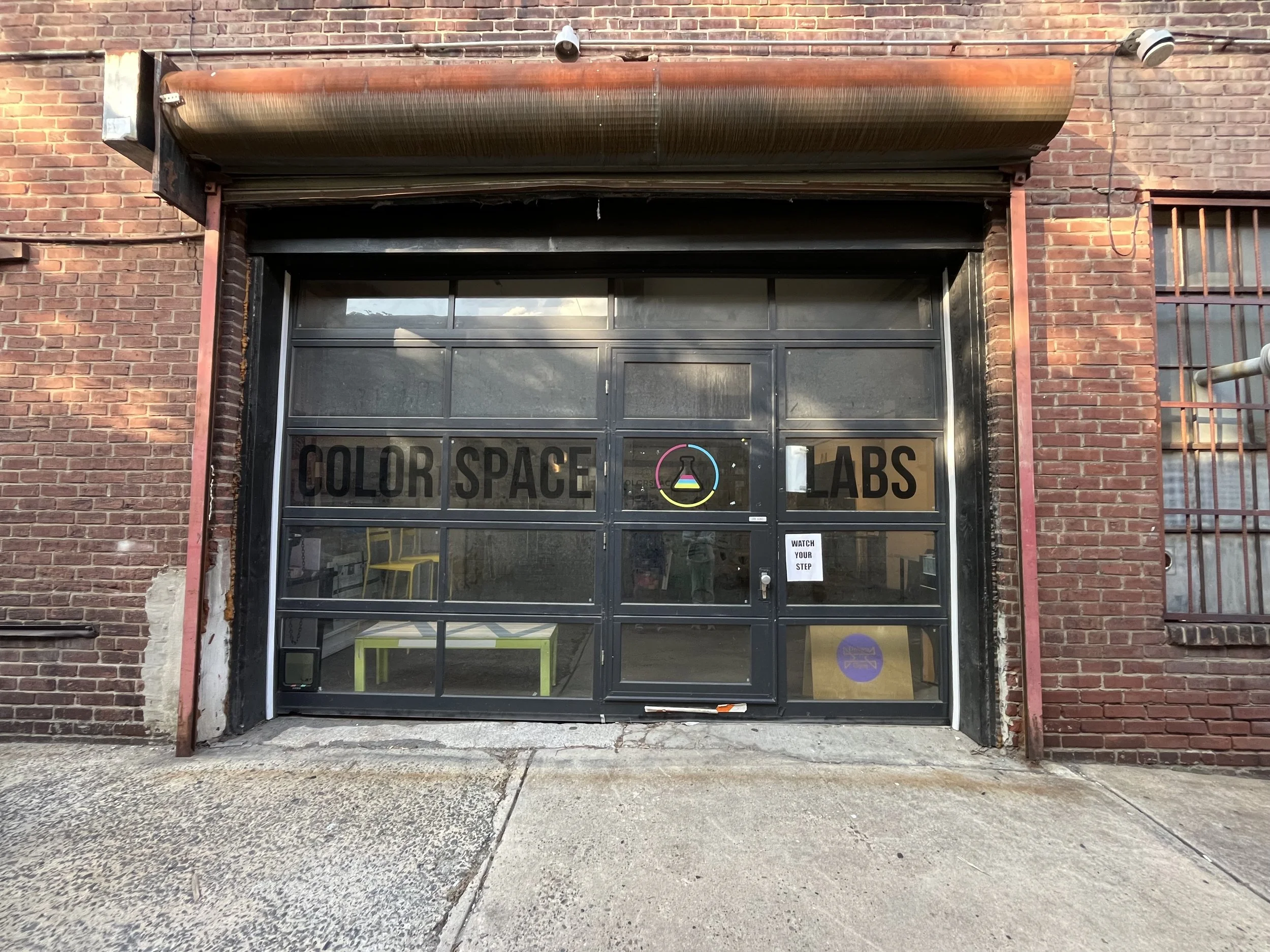 A view of glass garage doors with an entrance in it to enter a red brick industrial warehouse, with Color Space Labs in vinyl on the glass garage doors. Their logo, located at the door is a Narrow-Mouth Glass Flask with three lines of color.