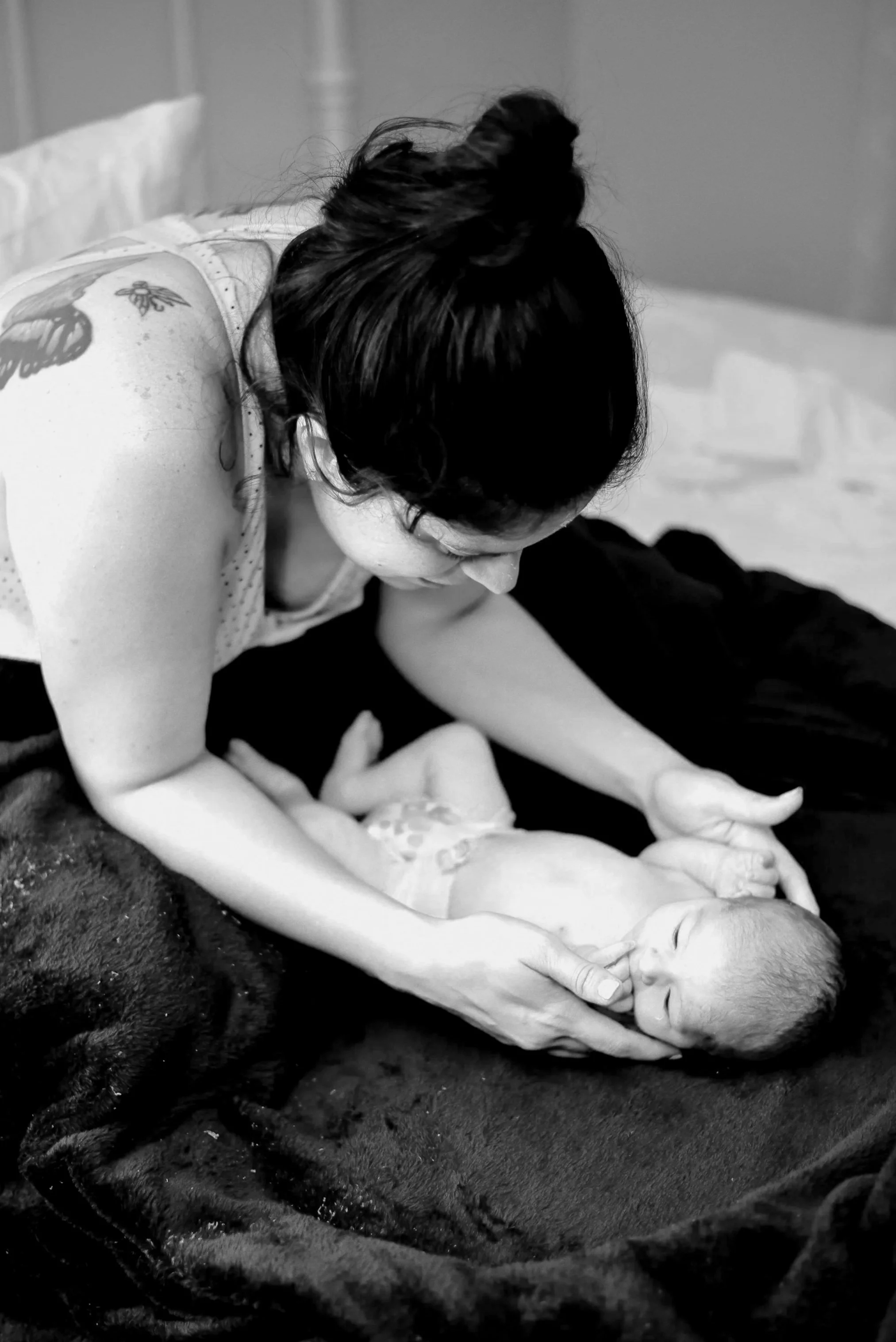 A woman with dark hair styled in a bun is lying down, gently cradling and caring for a newborn baby, both resting on a soft dark blanket in a grayscale photo.