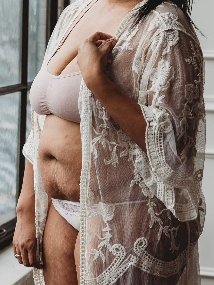 Woman in lace robe standing in soft light, embracing postpartum body and natural beauty after birth