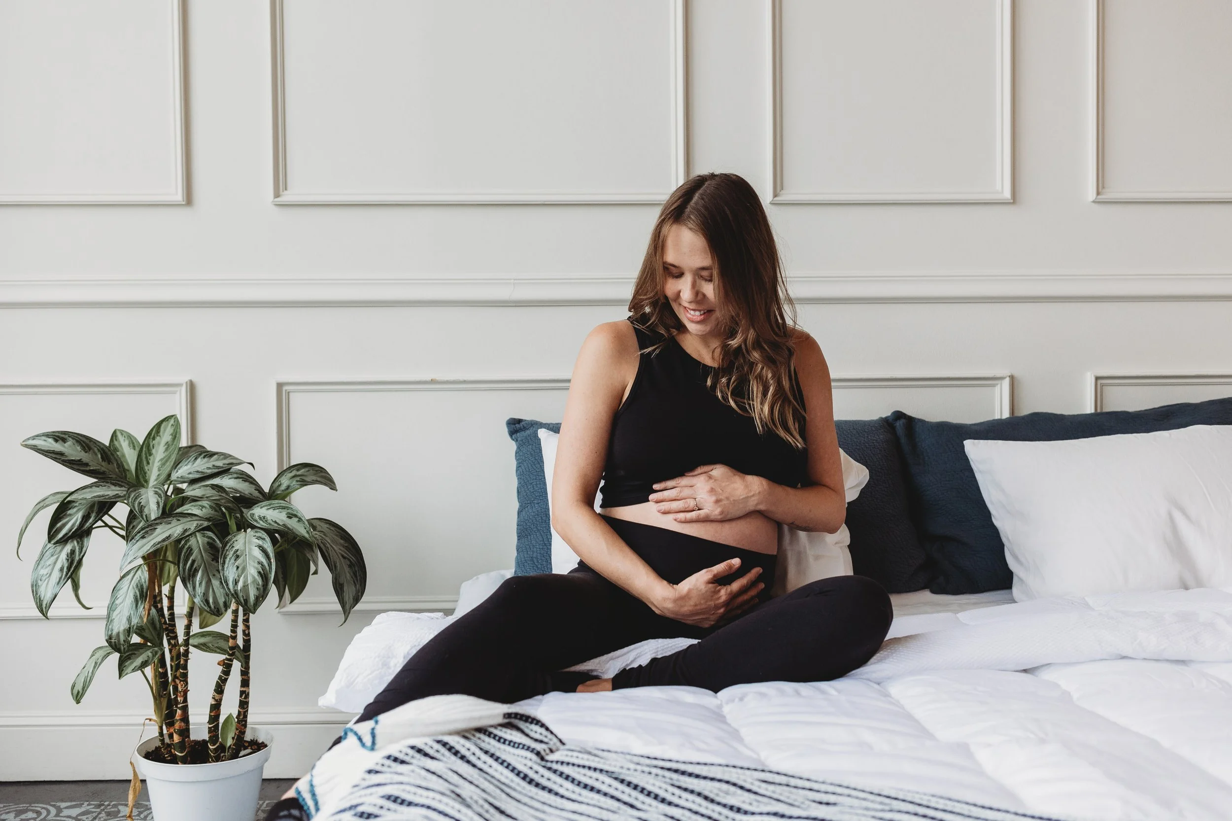 Pregnant woman sitting on bed holding belly, showing calm joy and anticipation during holistic pregnancy