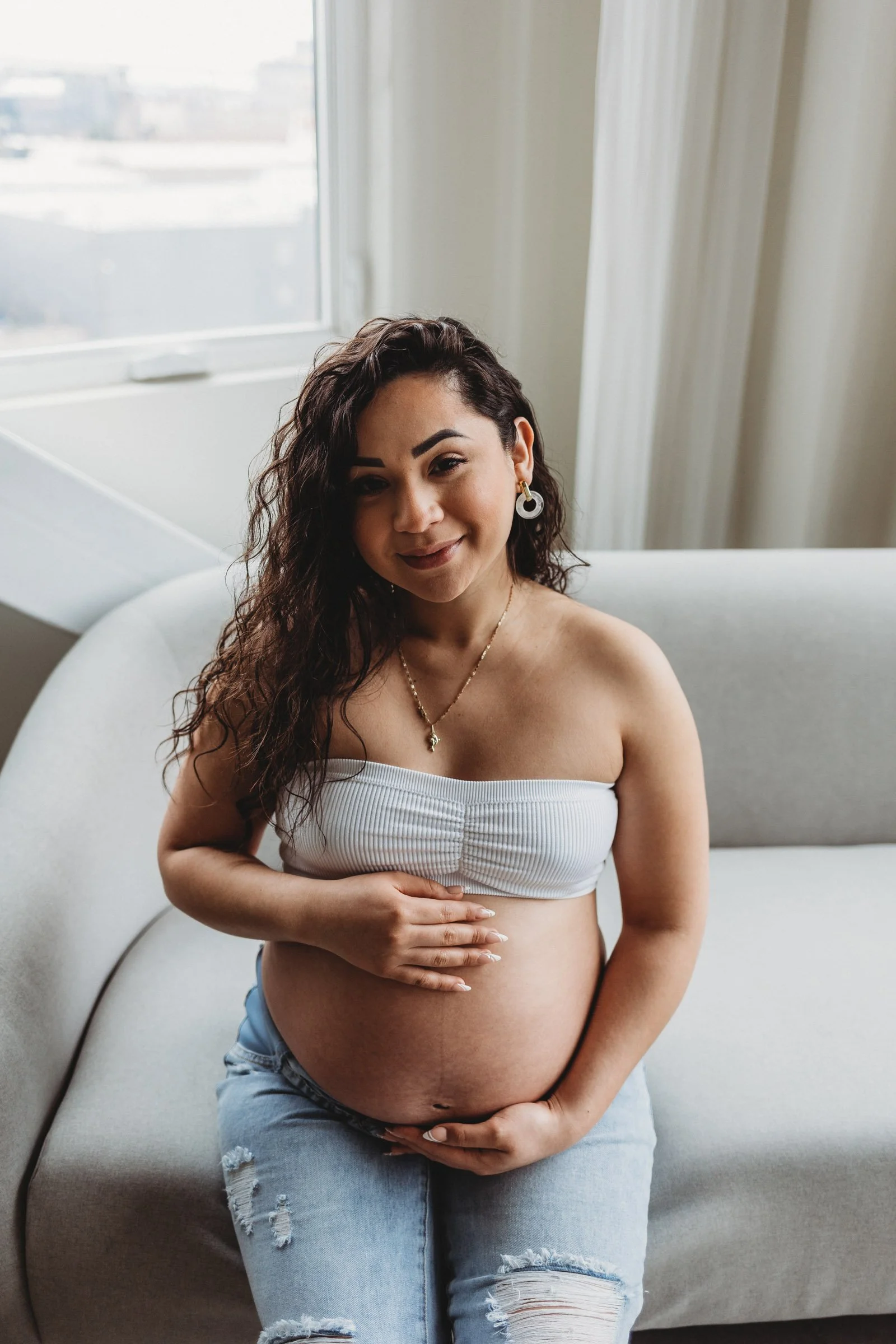Smiling pregnant woman in white top and jeans sitting on couch, showing joy and calm of holistic pregnancy