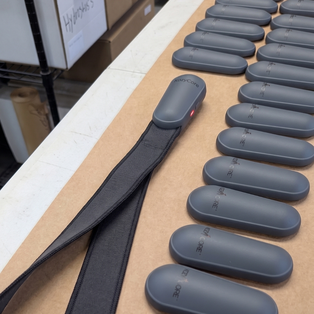 Finished KozyCore wearable device with fabric strap displayed next to a production line of manufactured enclosures
