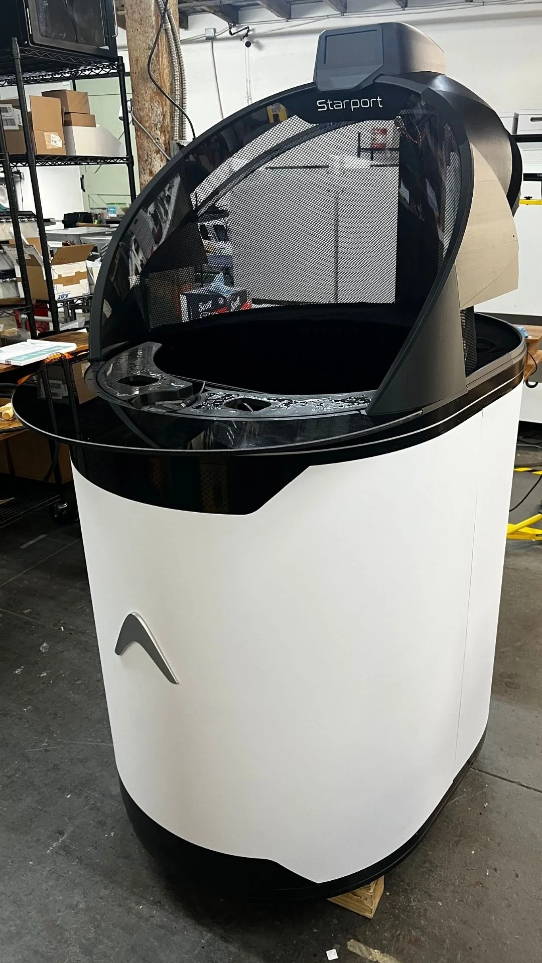A partially assembled Starport Explorer spatial portal prototype resting on a concrete floor in an industrial workshop. The device features a large white cylindrical base with a silver chevron logo and black trim at the bottom and top edges.