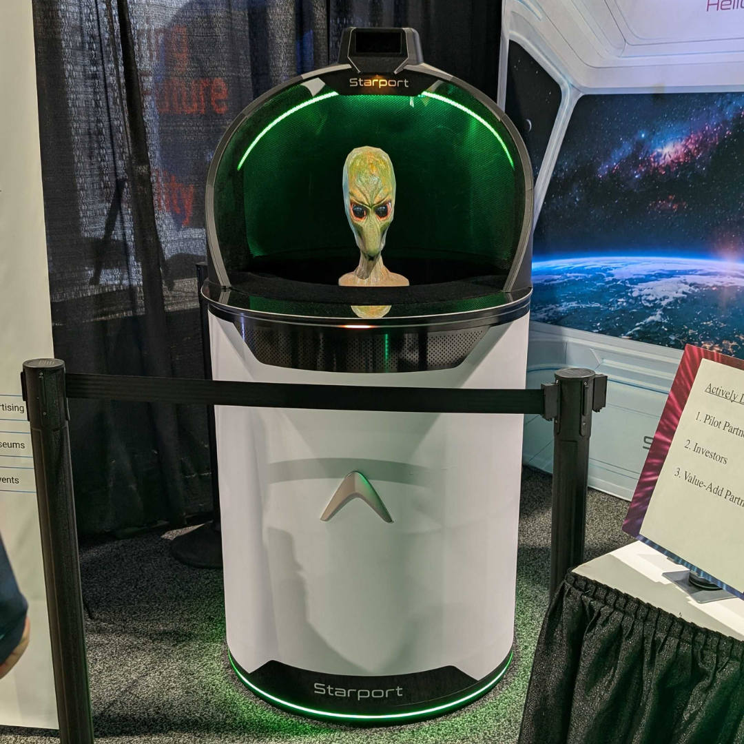 The Starport Explorer spatial portal on display at an exhibition, featuring a white cylindrical base and a glowing green dome rendering a 3D alien character.