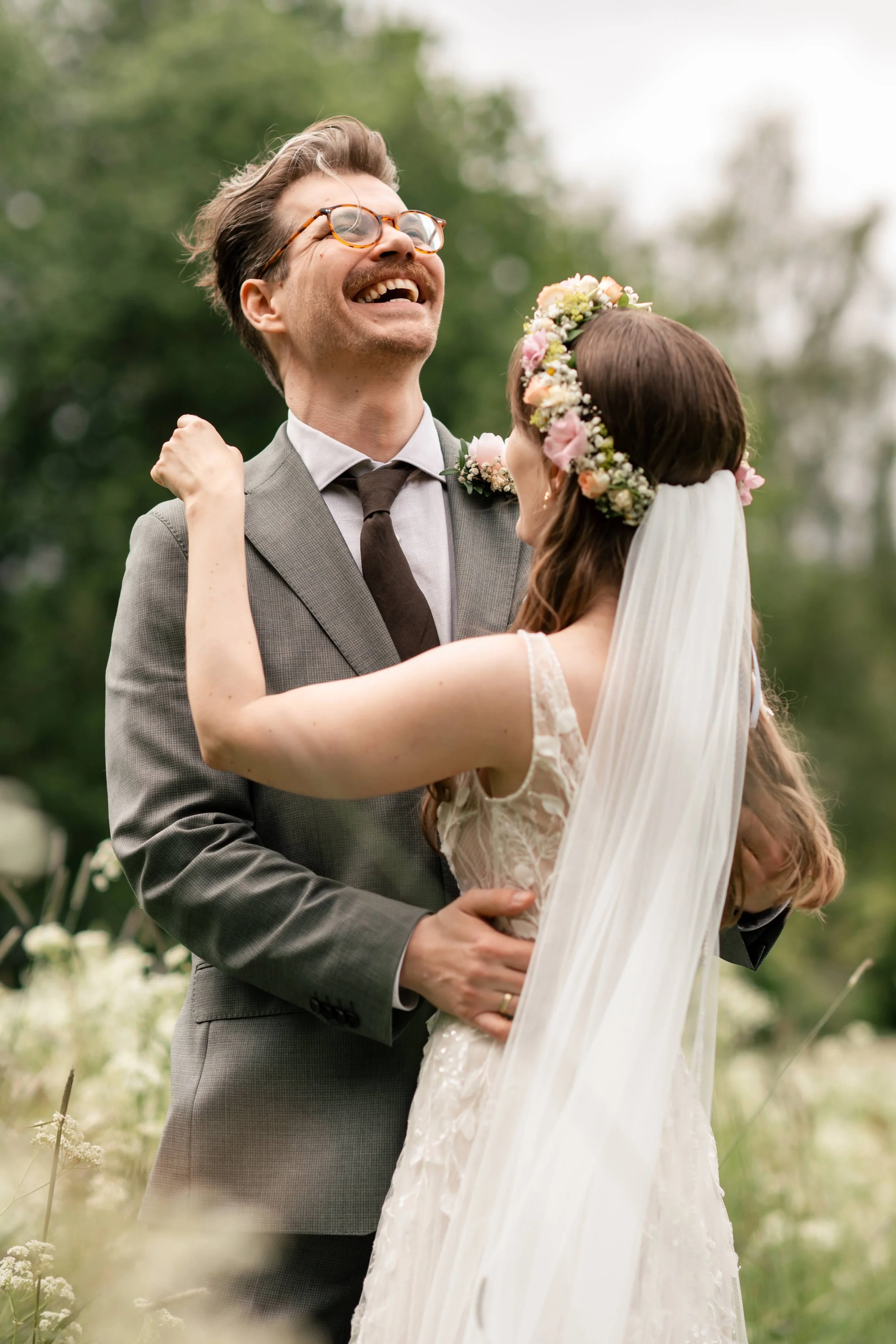 Et brudepar som smiler og koser seg i en grønn park, med blomster og trær i bakgrunnen.
