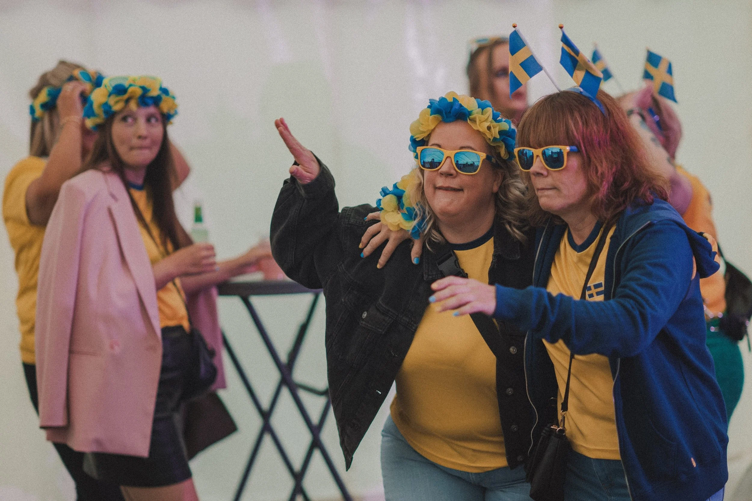 Grupp mennesker iført svenske flagg og farger som feirer, sammen med blomsterkranser og solbriller, i en innendørs setting.