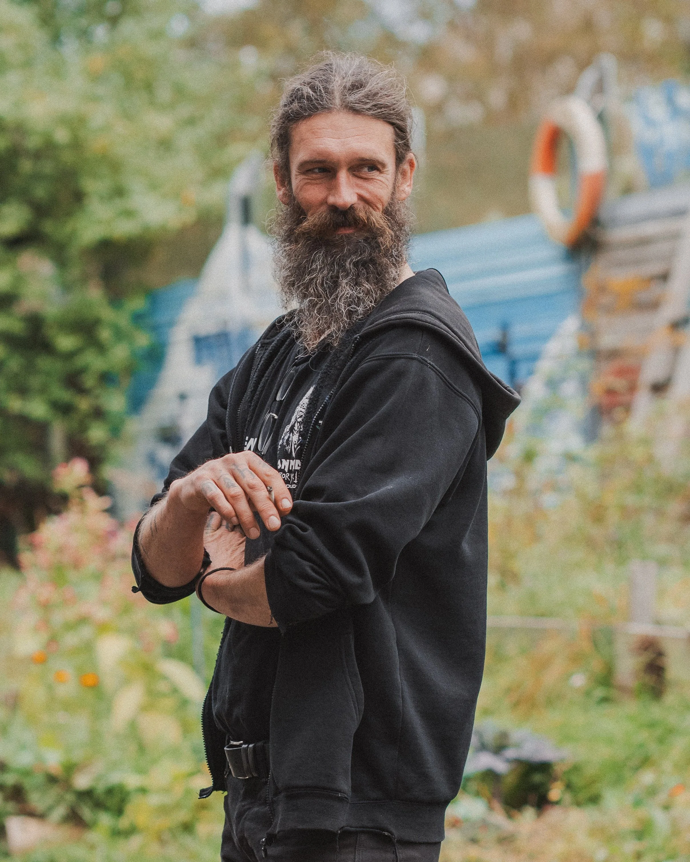 En mann med skallet hode og fullt skjegg, iført svart hettegenser, står utendørs med grønne trær i bakgrunnen.