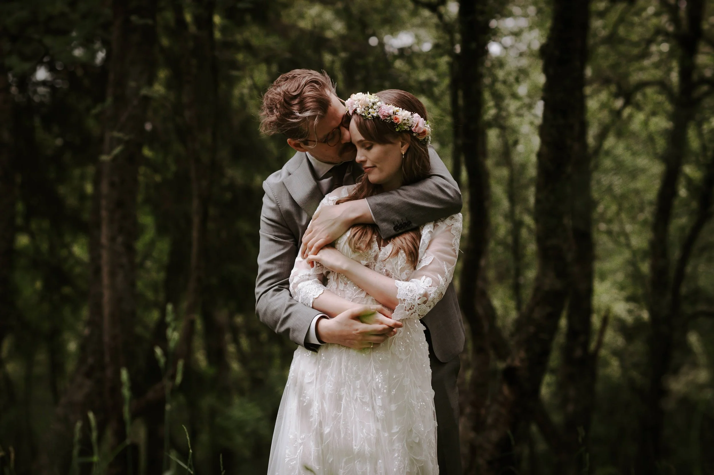 Et bryllupsportrett av et ungt par i en skog, der man ser et brylladsantrekk, blomsterhodepynt og omfavnelse.
