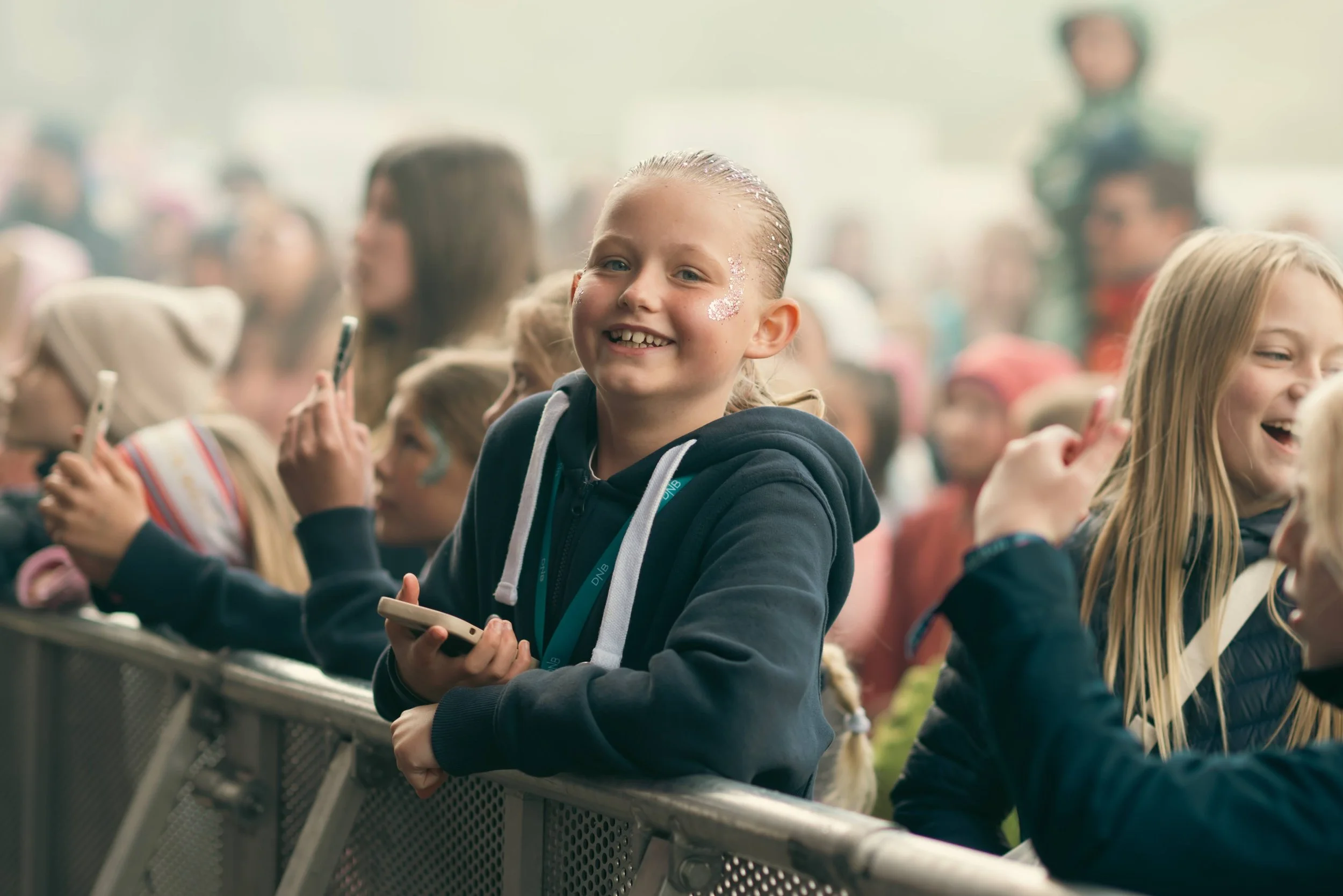 Ung jente smiler og har på seg en mørkeblå hettegenser, omgitt av en stor gruppe barn på en utendørs scenekonsert eller arrangement.