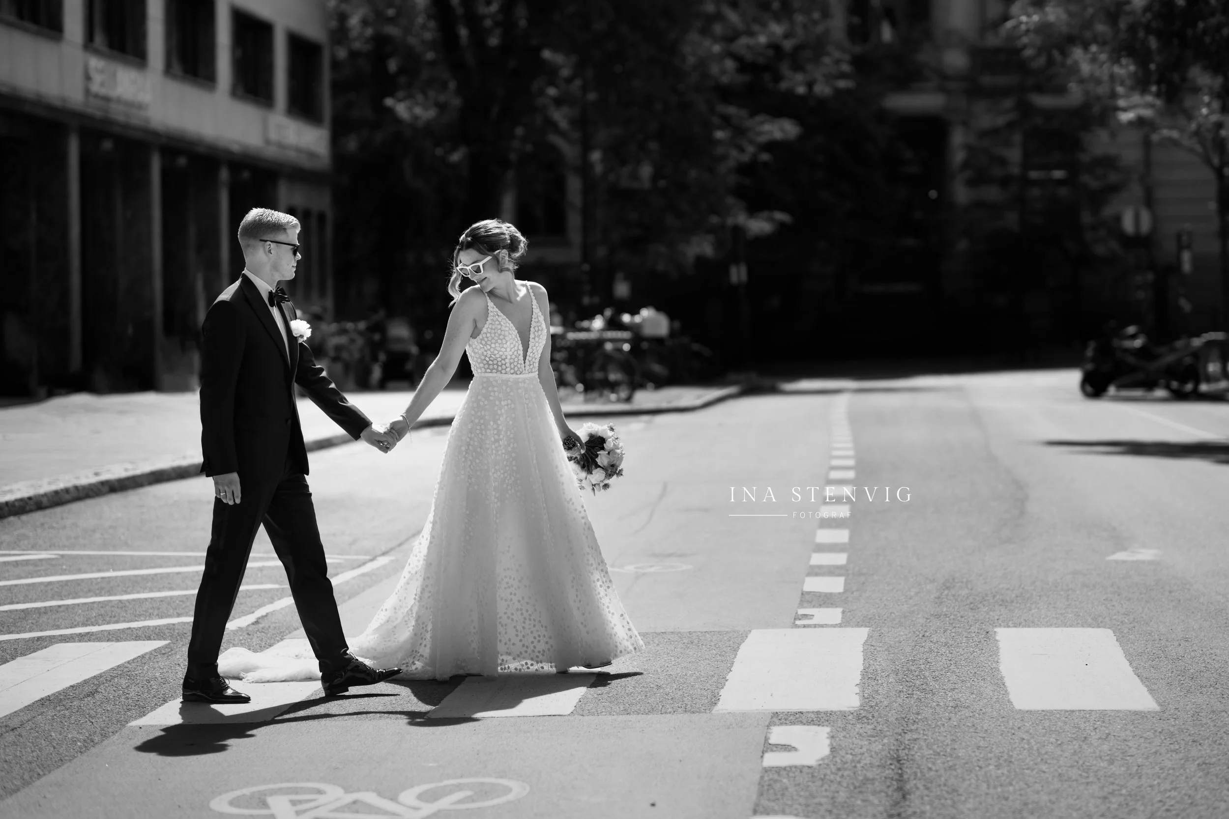 Et svart-hvitt bilde av et brudepar som holder hender mens de går over et fotgjengerfelt på en tom gate. Kvinnen har på seg en lang, hvit brudekjole med dyp utringning og bærer en bukett blomster, mens mannen har på seg en svart dress og slips.