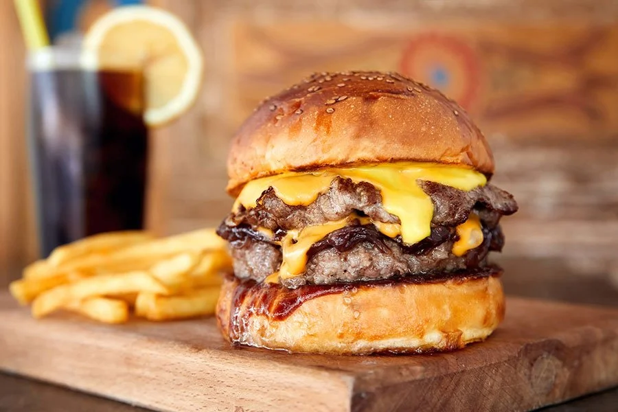 Close-up of a juicy cheeseburger with melted cheese, fries, and a soft drink on a wooden tray
