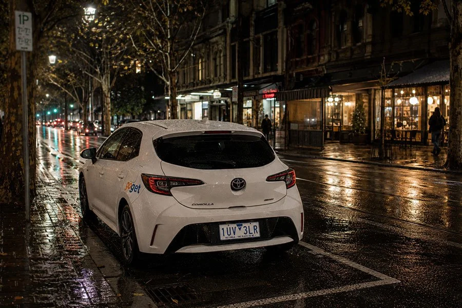 A white GoGet hatchback parked kerbside on a wet Melbourne street at night, with shopfront lights reflected in the rain-slicked road.