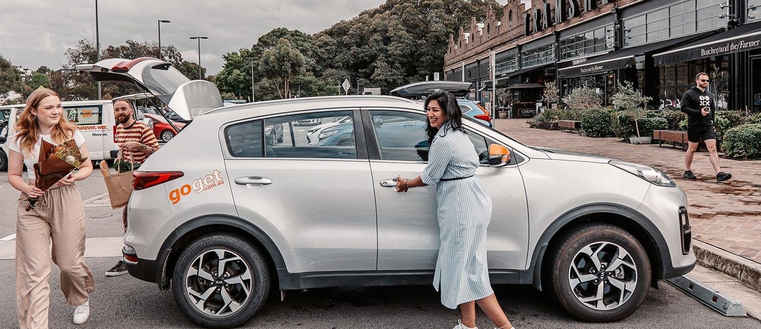 A smiling woman opens the door of a GoGet carshare SUV, with 2 passengers unloading food and flowers from the boot
