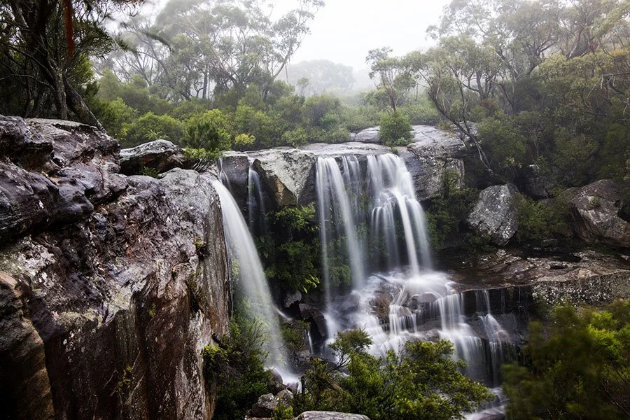 Wide waterfall cascading into a deep shaded swimming hole beneath a sandstone cliff in a national park south of Sydney