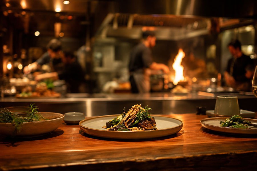 A plated dish on a timber chef's counter in a warmly lit open restaurant kitchen, with chefs and a wood fire softly blurred behind.