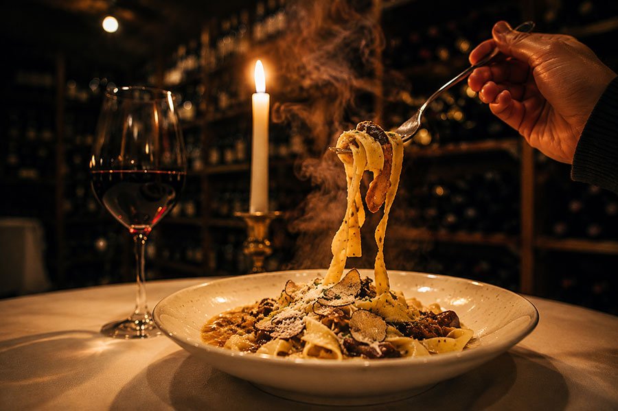 A fork lifting steaming ribbons of pappardelle from a bowl of ragù topped with shaved truffle and parmesan, beside a lit candle and a glass of red wine, with a wall of racked wine bottles blurred in the darkness behind.