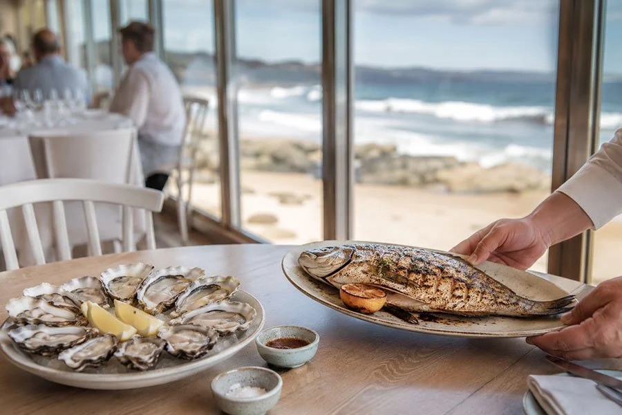 A waiter placing a whole grilled fish beside a platter of freshly shucked oysters at a beachfront restaurant table, with surf visible through floor-to-ceiling windows.