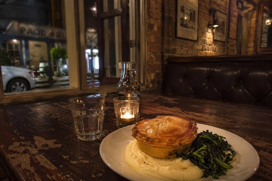 A golden pastry pie served on mashed potato with sautéed greens, lit by candlelight on a timber table in a heritage brick pub in Paddington, Brisbane.
