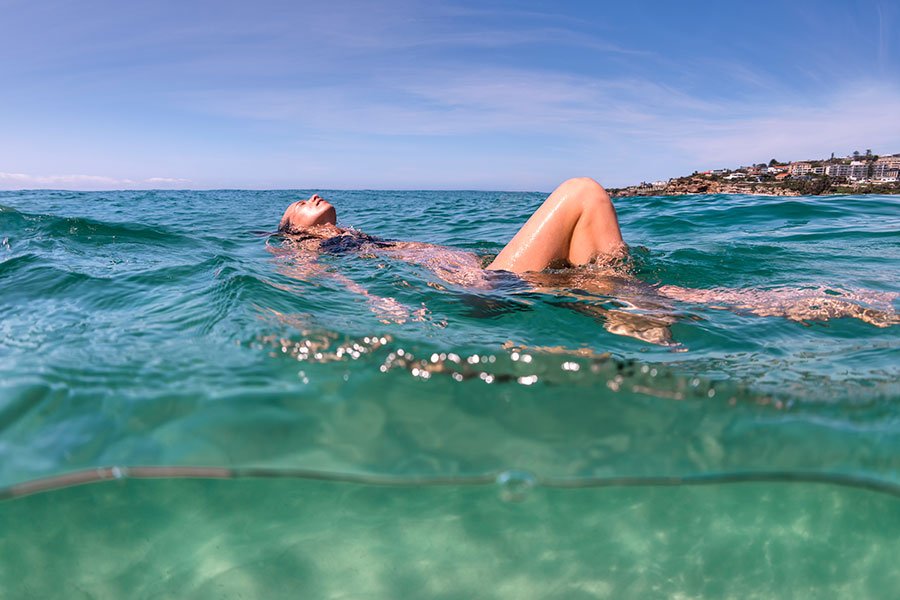 Woman floating in clear turquoise ocean water on a sunny Sydney summer morning, coastal suburb visible in the background
