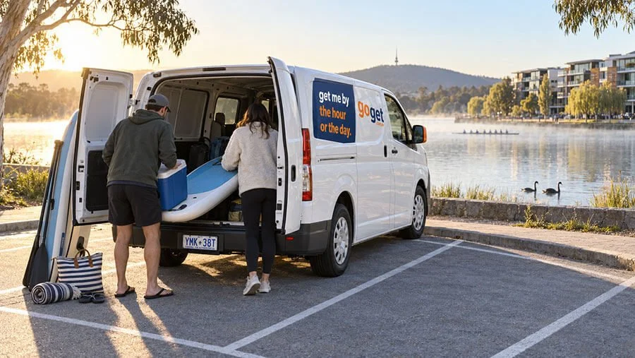 Two people loading paddleboards and an esky into a white GoGet van parked beside a misty lake at sunrise, with black swans, rowers and lakeside apartments behind.