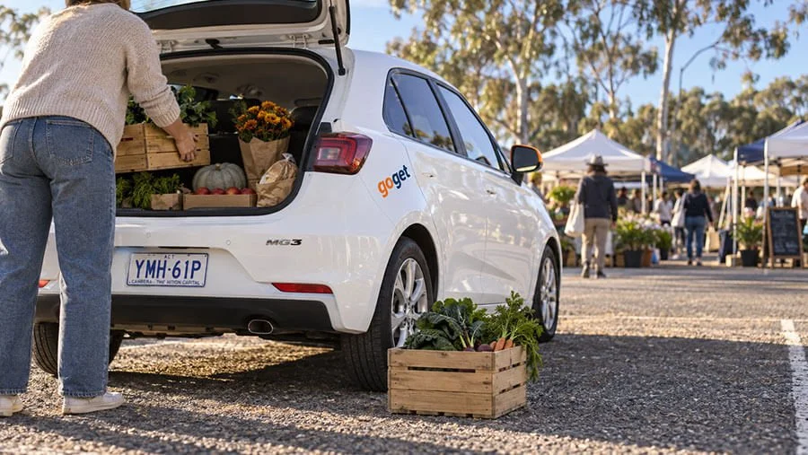 Person loading a timber crate of vegetables into the open boot of a white GoGet hatchback with an ACT number plate at a farmers market car park lined with gum trees.