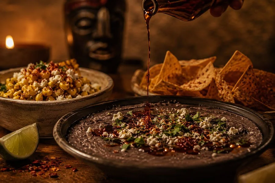 Chipotle oil poured over a bowl of smoked black bean dip topped with crumbled cheese and coriander, beside esquites, corn totopos and a lime wedge.