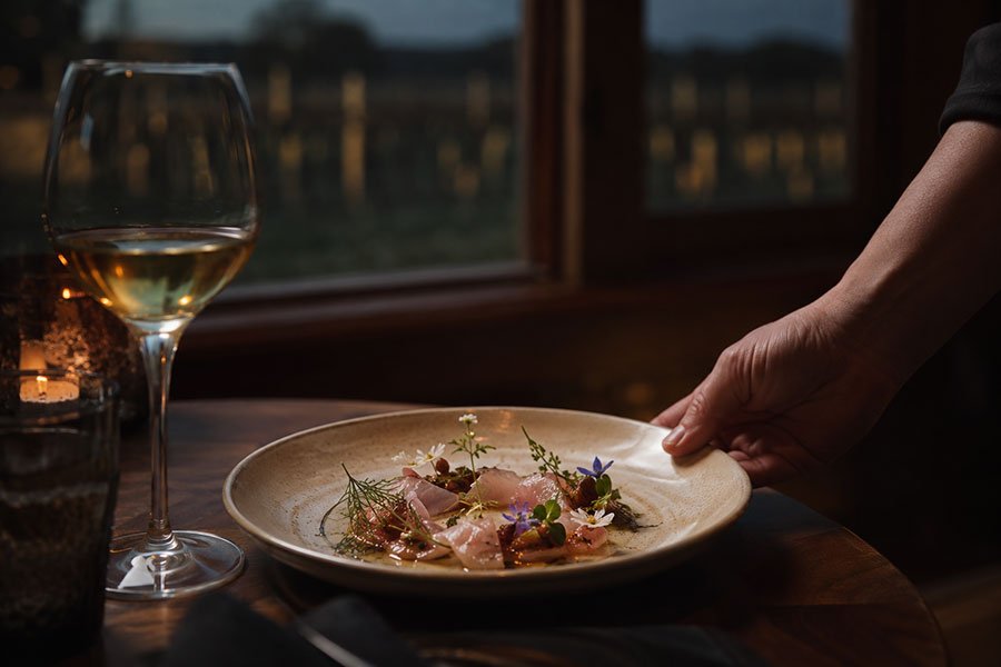 A delicately plated dish garnished with edible flowers served by candlelight, with a glass of white wine and dusk vineyard views through the window.