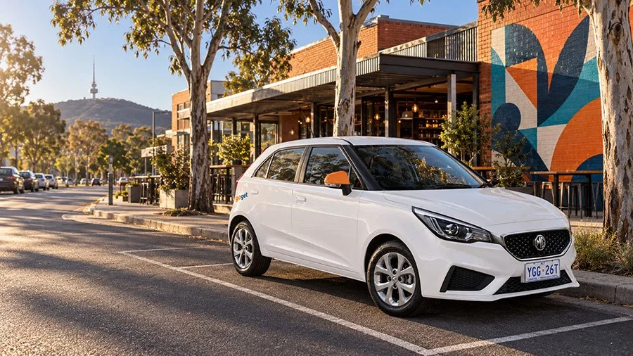 White GoGet carshare hatchback with orange side mirrors parked in a marked bay on a tree-lined Canberra street at sunset, with a communications tower on a distant hill and a colourful mural behind.