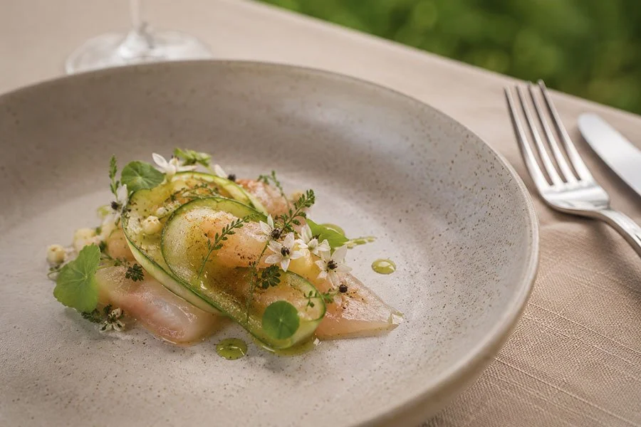 A delicately plated seafood dish garnished with herbs and edible flowers on a textured ceramic plate, with soft green lawn blurred in the background.