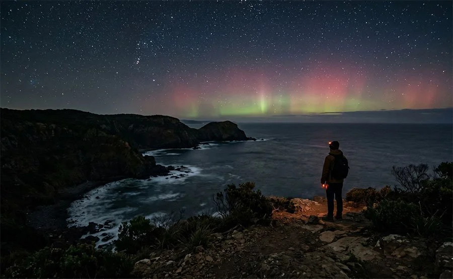 Stars and a faint aurora glow above a rugged southern coastline with dark cliffs and Bass Strait visible below.