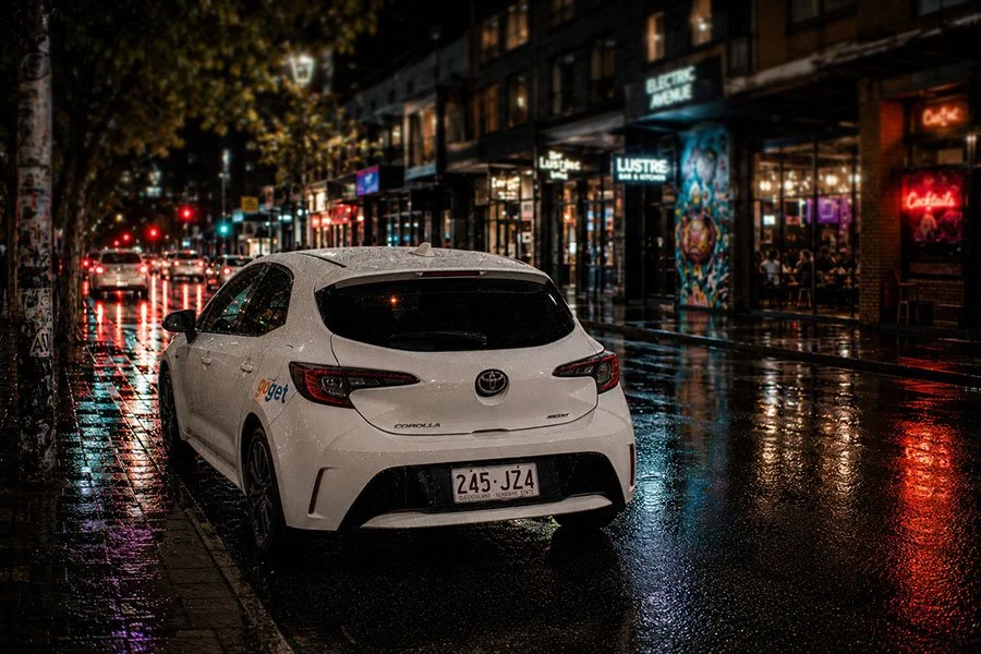 A white GoGet Toyota Corolla parked on a wet inner-Brisbane street at night, with lit shopfronts and traffic lights reflected on the road.