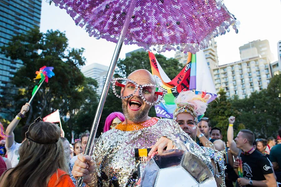 Joyful festival-goer in a full sequin costume and oversized diamante glasses holding a glittering umbrella at an outdoor pride celebration in Sydney's CBD