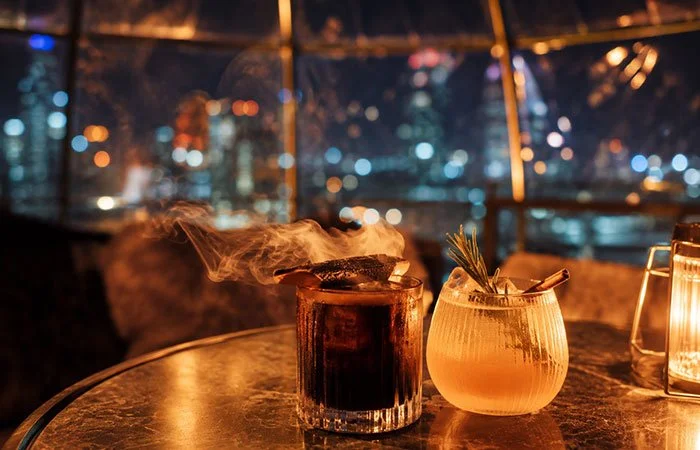 A glowing transparent dining igloo on a terrace at night, with the Brisbane city skyline lit up behind it.