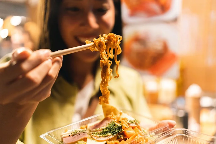 Woman enjoying street food at a night market, seated outdoors with chopsticks and warm lighting — perfect winter foodie vibes