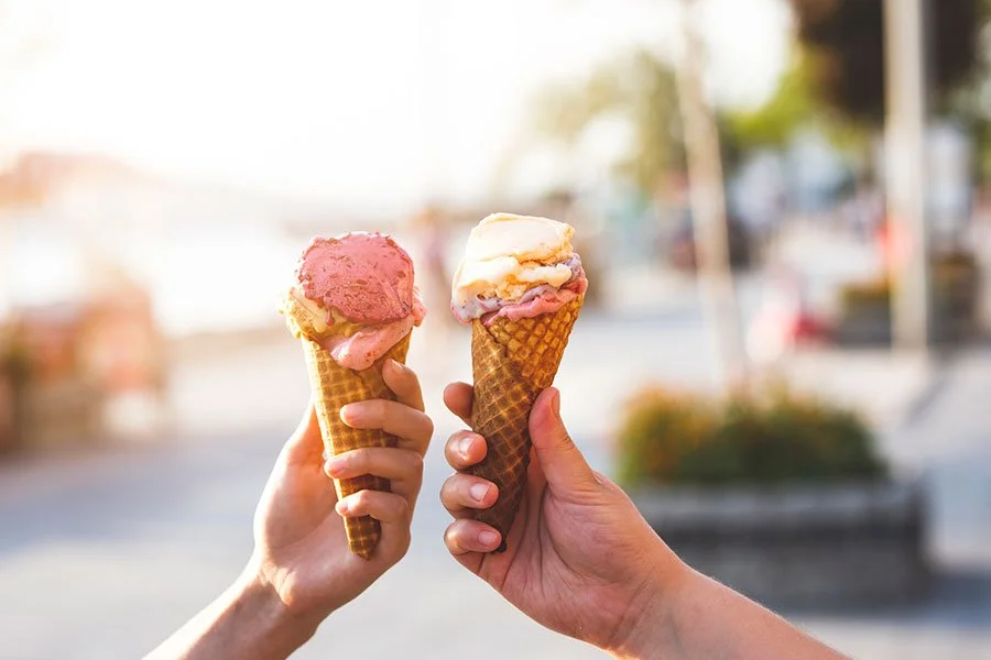 Two hands holding waffle cones filled with creamy gelato and colourful toppings at an outdoor dessert stall