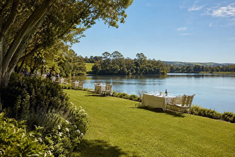White-clothed tables set on a lawn beside a still lake, surrounded by garden greenery under a clear sky.