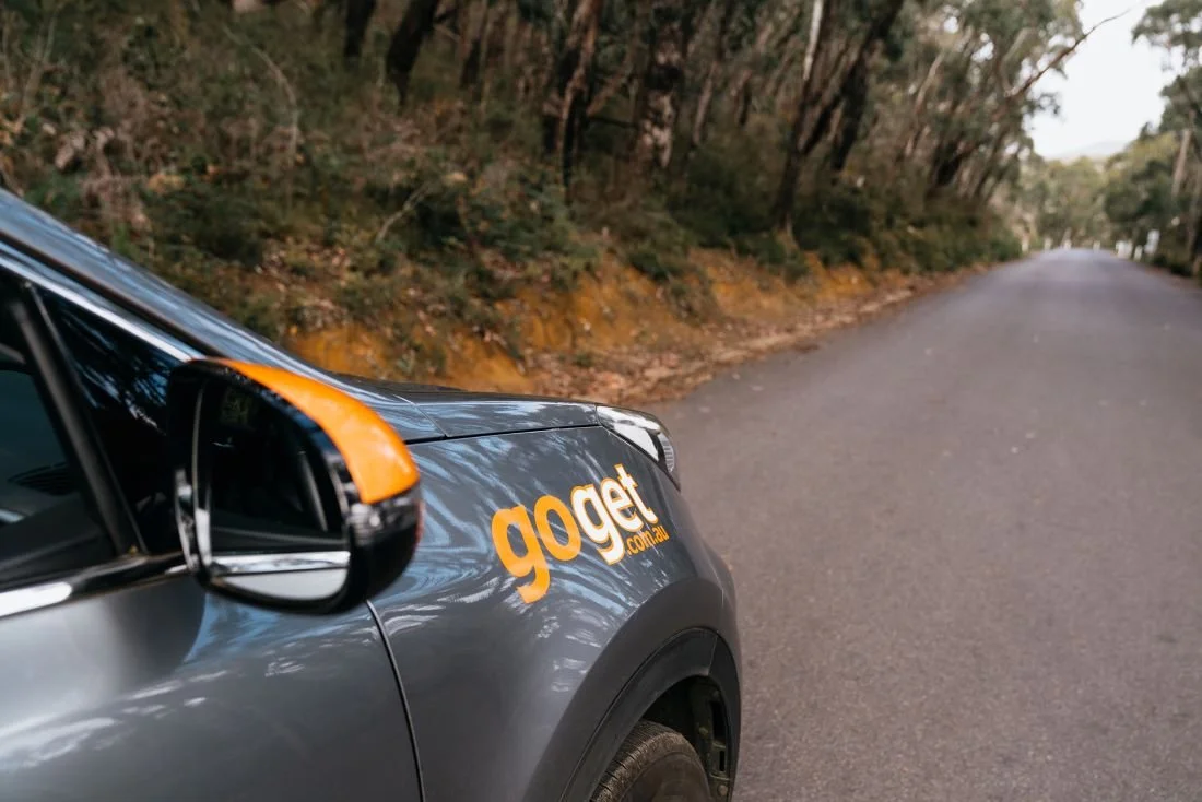 The front of a grey GoGet car with orange mirror detailing parked on a quiet country road, tall eucalypts arching overhead.