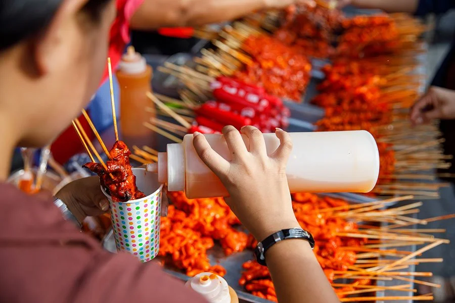 Vendor pouring sauce over grilled Filipino barbecue skewers at a multicultural outdoor food stall