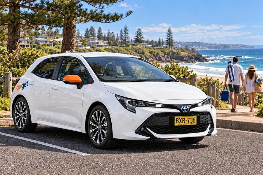 A white GoGet hatchback with an orange wing mirror parked in a beachside car park, as two people carrying a cooler bag and a beach umbrella walk towards the beach past Norfolk Island pines.