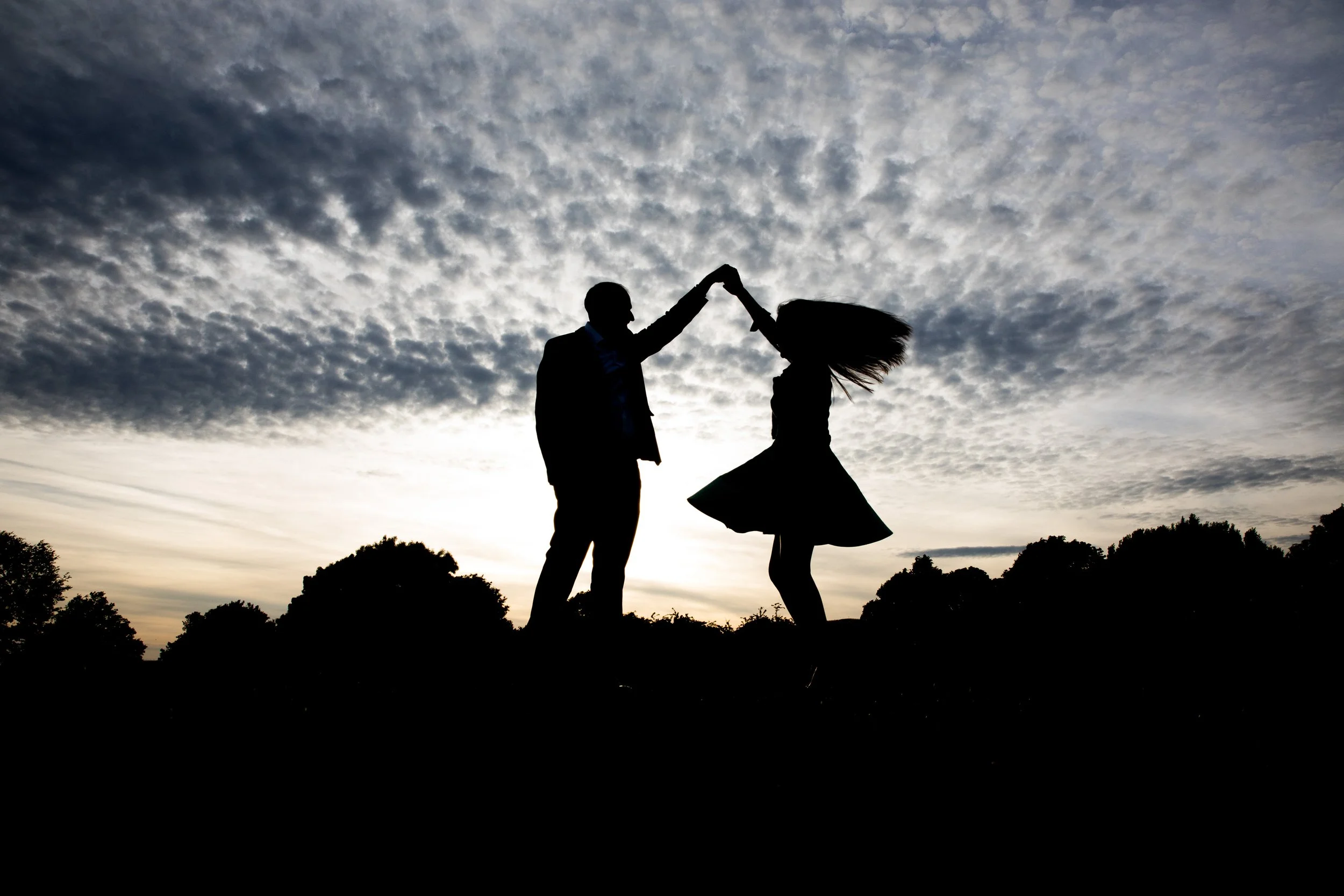 A silhouette of a man and woman dancing outdoors against a cloudy sky at sunset.